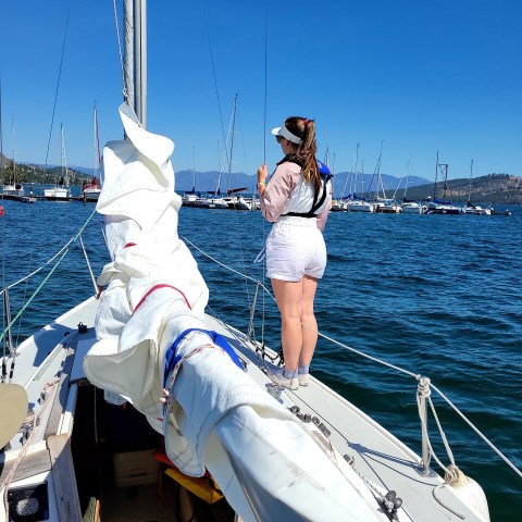 Person standing on a sailing boat deck, looking at water and distant sailboats on a clear day.
