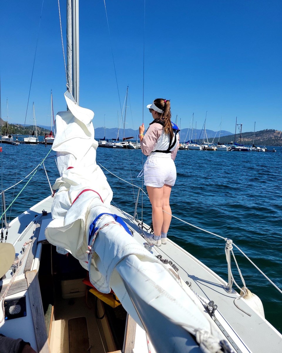 Person standing on a sailing boat deck, looking at water and distant sailboats on a clear day.