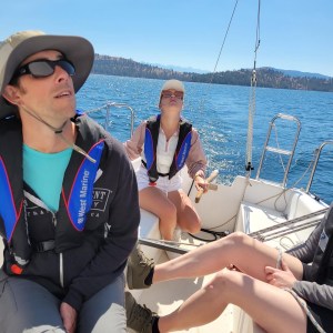 Three people wearing life vests on a sailboat, with a lake and forested hills in the background.