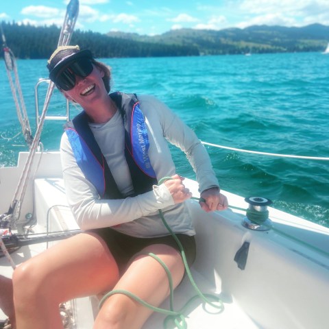 Person smiling on a sailboat, wearing a life jacket and sunglasses, pulling on a rope.