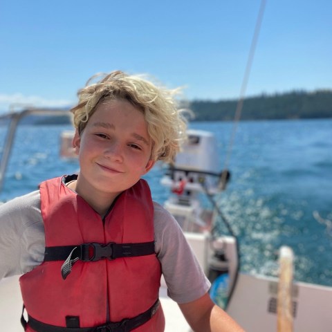 Child in red life jacket on a sailboat with blue water and forested shore in background.