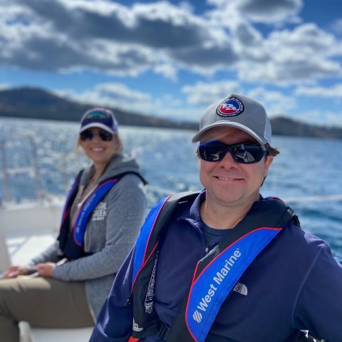 Two people in life jackets on a boat, smiling under a partly cloudy sky.