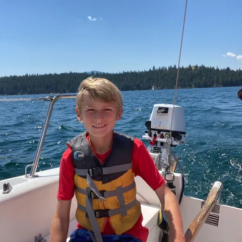 Smiling child in a life jacket on a sailboat on a sunny day, with forested shore in the background.