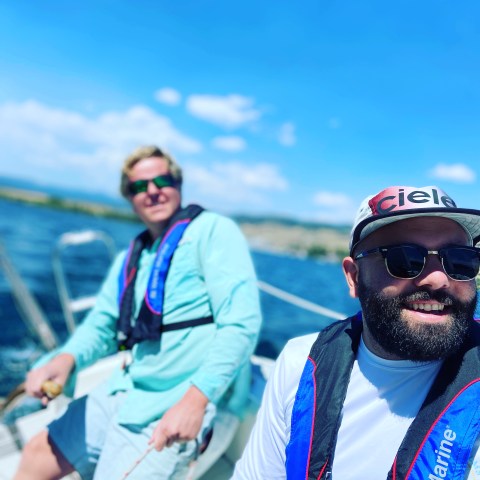 Two men sailing on a boat under a clear blue sky, wearing life jackets and sunglasses.