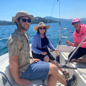 Three people smiling on a sailboat with a lake and mountains in the background.