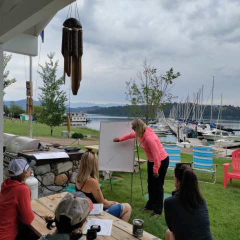 Outdoor workshop by a lake with a woman writing on a whiteboard and people seated nearby.