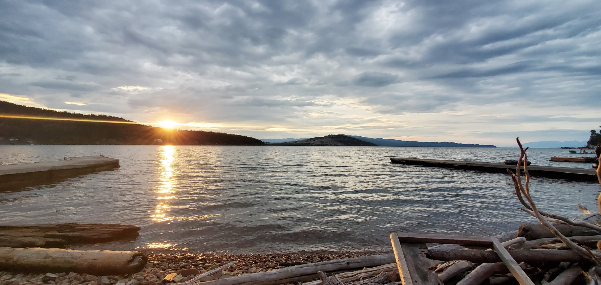 Sunset over a lake with docks, driftwood on pebbled shore, and cloudy sky.