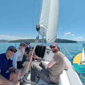Four people sailing on a boat with a logo on the water, clear sky.