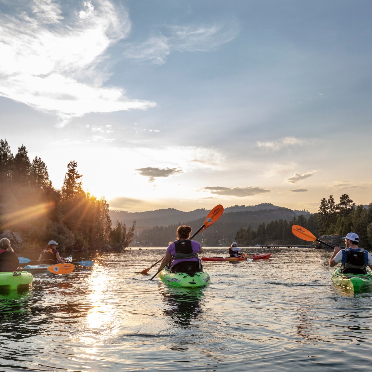Group of kayakers paddling on a lake at sunset with trees and mountains in the background.